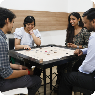 Four adults sit around a carrom board in a bright room, actively setting up the game pieces and chatting.