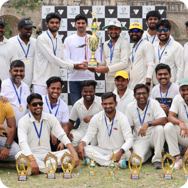 Cricket team posing for a trophy photo on a grassy field, with central players holding a large trophy and wearing medals; teammates stand and kneel around them, small trophies lined up in front.