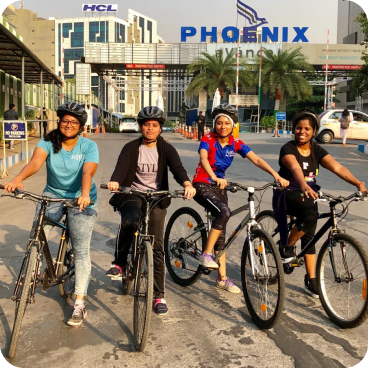 Four children wearing helmets ride their bicycles side by side on a sunny city street, with palm trees and a large Phoenix sign in the background.
