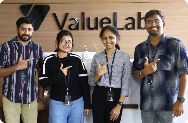 Four coworkers stand together in front of a ValueLabs sign, smiling and pointing at the logo.