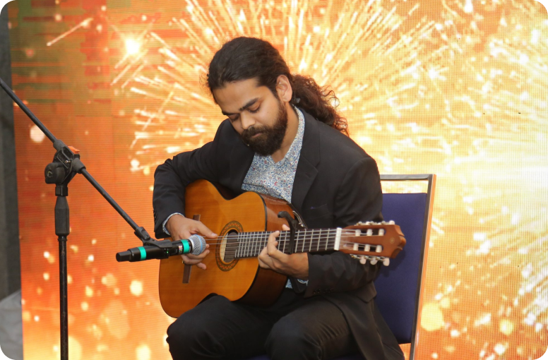 Man with long dark hair and beard, wearing a blazer, sits on a chair playing an acoustic guitar on stage as a microphone is set up nearby and a bright orange spark backdrop glows behind him.